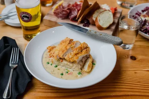 Plate of breaded fried chicken with green peas and creamy gravy, served with bread on a rustic wooden table, alongside a glass of beer, a knife on the plate, and a black napkin with a fork.