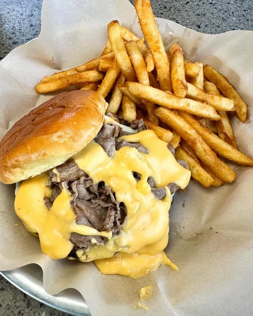 French fries and a cheeseburger with sliced beef and melted cheese in a bun on a metal tray lined with parchment paper.