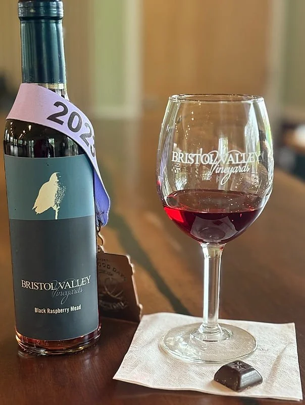 A bottle of Bristol Valley Vineyards Black Raspberry Mead with a wine glass filled with red wine on a table, and a piece of chocolate on a napkin.
