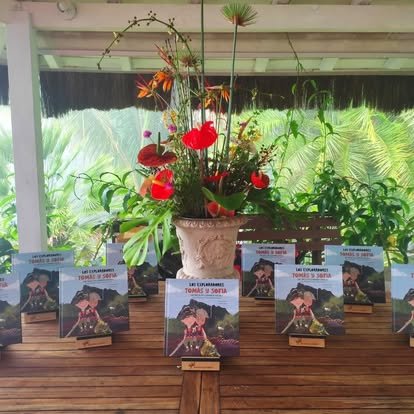 A flower arrangement with red and orange flowers in a large beige pot, accompanied by several copies of a book titled 'Tomas y Sofia' displayed on small stands on a wooden surface.
