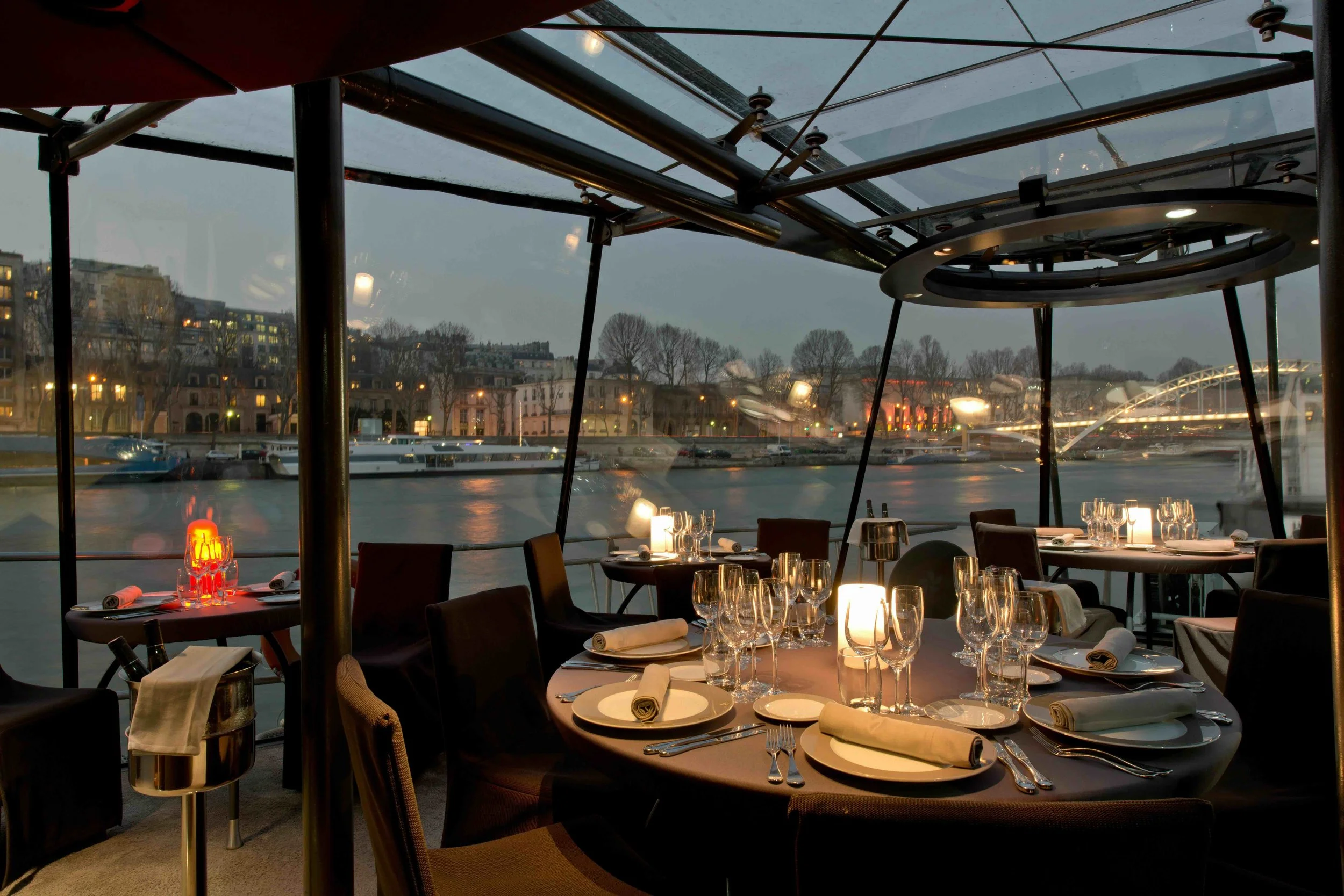 Indoor restaurant with tables set for dinner, overlooking a river and cityscape at dusk, with reflections on the water and city lights.