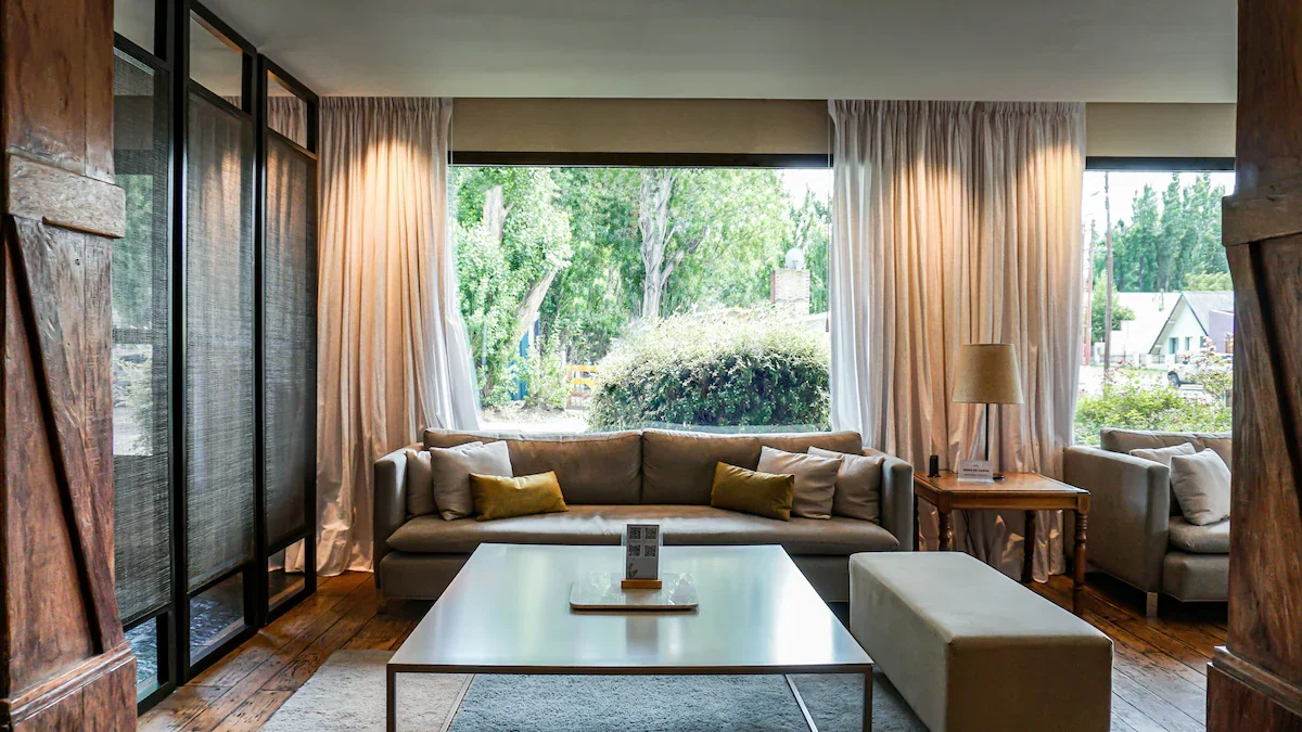Living room with large window showing green trees outside, beige sofa with pillows, wooden side table with a lamp, white coffee table, white bench, and curtains.