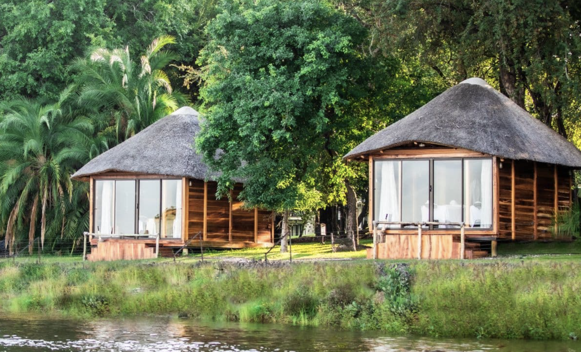 Two wooden huts with thatched roofs by a river, surrounded by lush green trees and foliage.