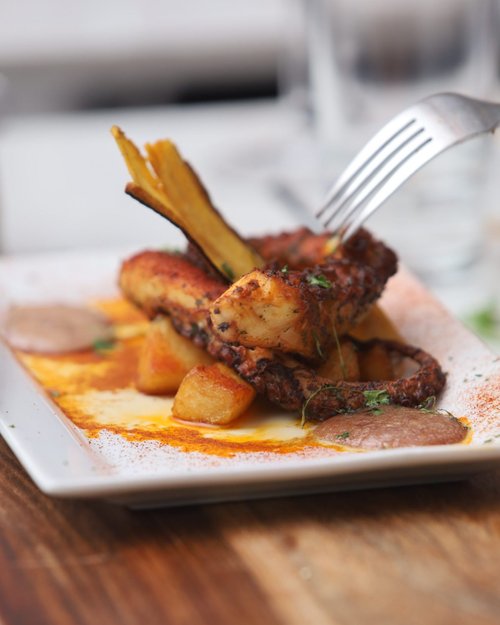 Close-up of a plated dish featuring grilled chicken with a spicy red sauce, golden roasted potatoes, fried plantains, and garnished herbs on a white rectangular plate.