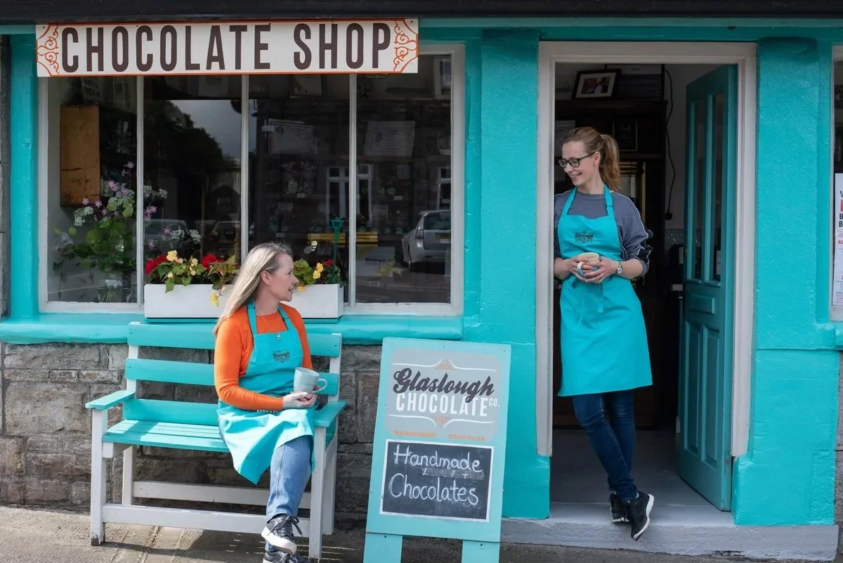 Outside a blue chocolate shop with a sign that reads "Chocolate Shop" and a chalkboard sign that says "Handmade Chocolates." A woman is sitting on a light blue bench with a teal apron, holding a cup, while another woman in a teal apron and glasses st