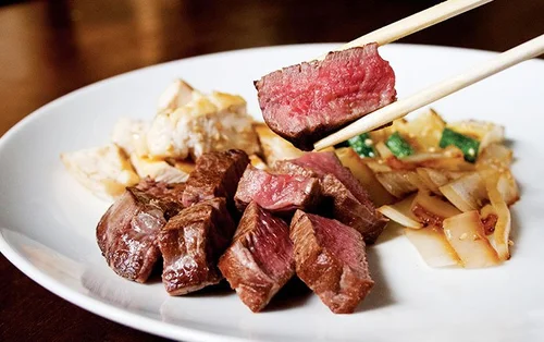 Slices of cooked steak with chopsticks holding a piece, served with noodles and vegetables on a white plate.