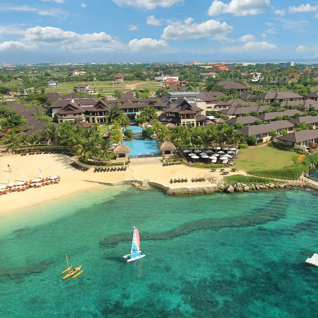 Aerial view of a luxurious tropical resort with a beach, pool, palm trees, cabanas, and sailboats in turquoise waters.