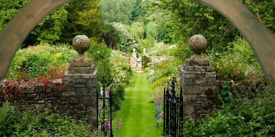 A garden viewed through an arched stone gate with iron gates, lush greenery, and colorful flowers.