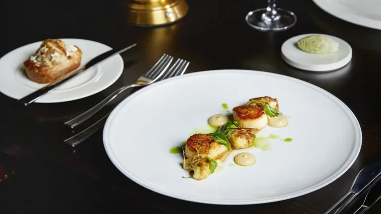 A plate with seared scallops garnished with microgreens and sauce, a dessert pastry on a small plate, and a small dish of green condiment on a dark table with silverware and wine glass in the background.