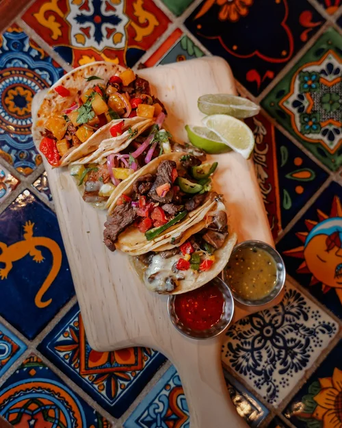 Three beef tacos with cheese, vegetables, and salsa on a wooden serving board, garnished with lime wedges and accompanied by two small bowls of salsa and sauce, set on a colorful tiled table.