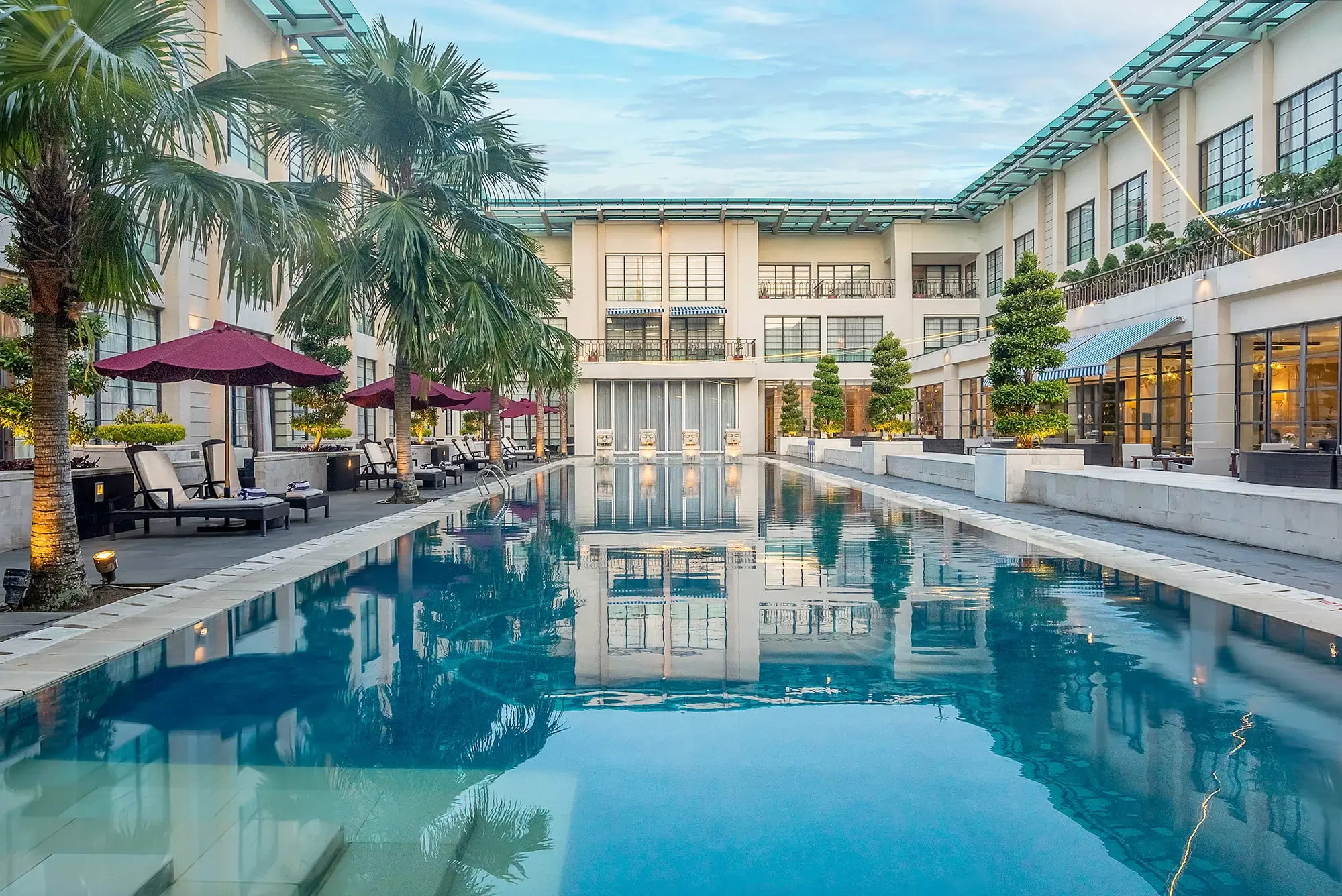 A modern hotel courtyard with a swimming pool, lounge chairs, umbrellas, and lush greenery, surrounded by a multi-story building with large windows and balconies, under a cloudy sky.