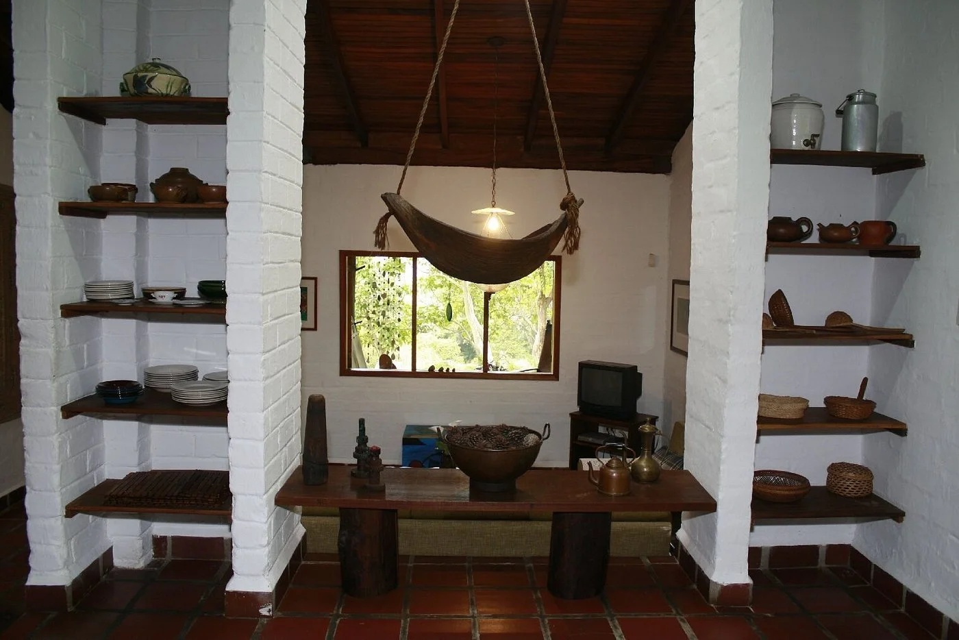 Living room with white brick shelves holding pottery and bowls, a window with green trees outside, a wooden table with decorative items, and a hammock hanging from the ceiling over the window.