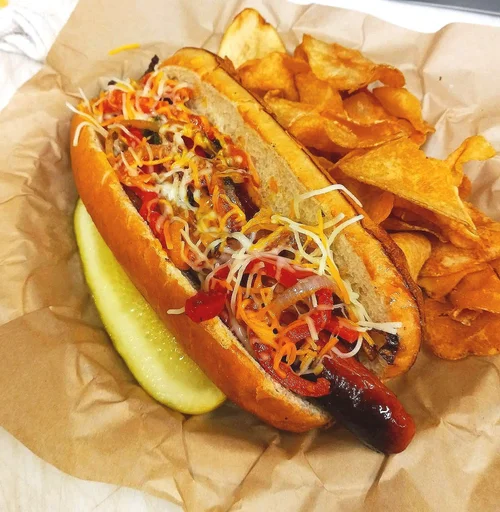 Hot dog topped with chili, shredded cheese, and diced peppers, served with potato chips and a lemon wedge.