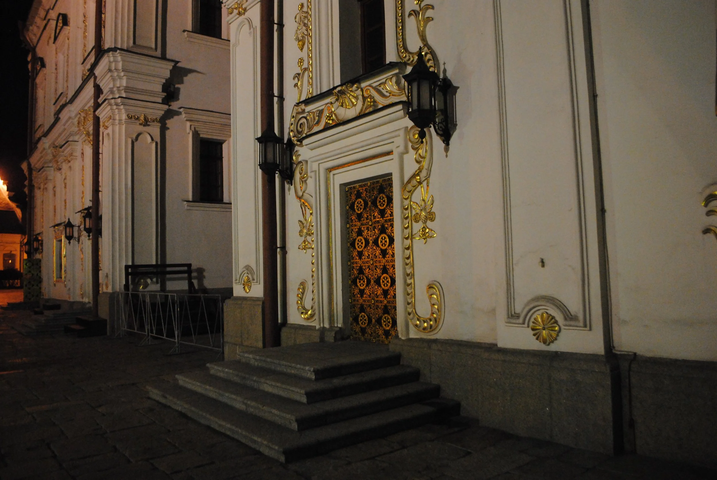 Night view of an ornate, white and gold decorated building with a black door backed by steps. The building features intricate gold leaf accents and decorative black lanterns on the exterior.