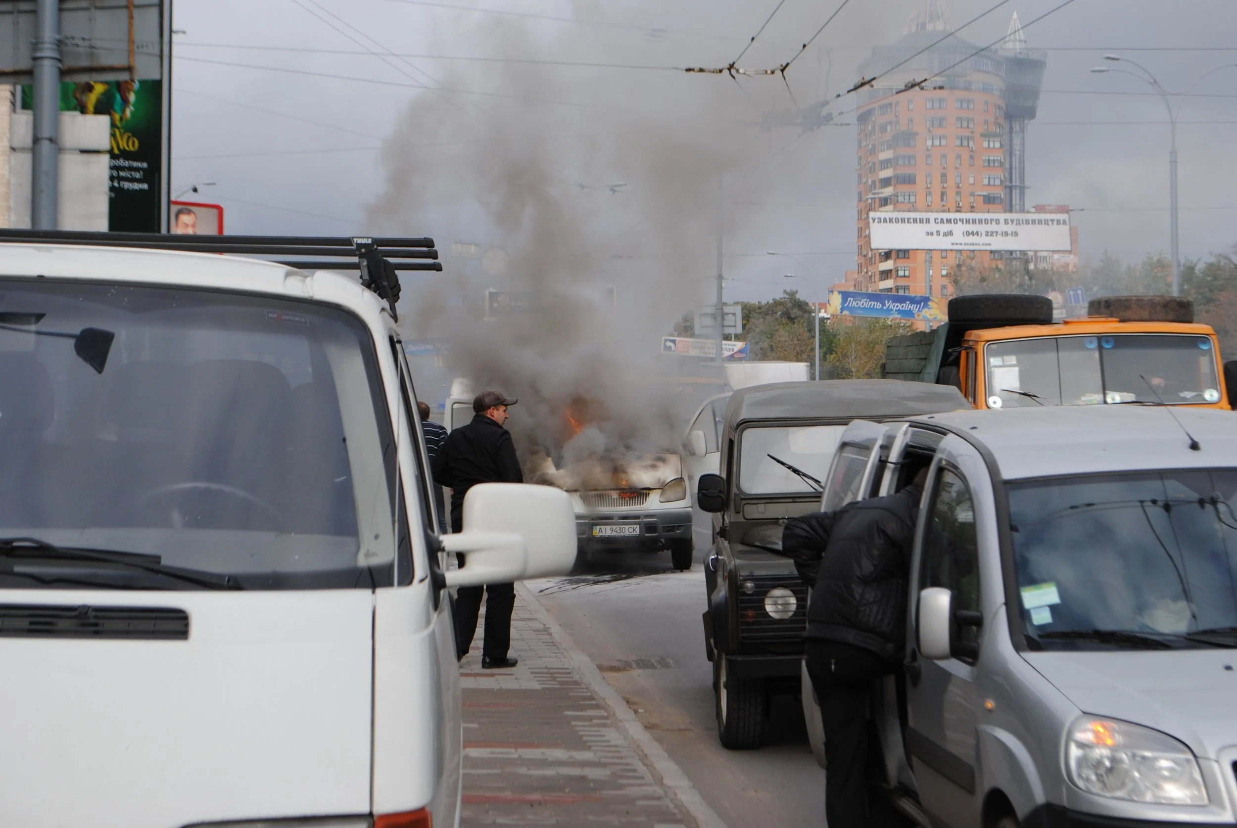 A busy city street with multiple vehicles and a fire causing smoke, with a person standing near the fire and smoke rising in the background. Kiev November 2011, Marina Parrish ©