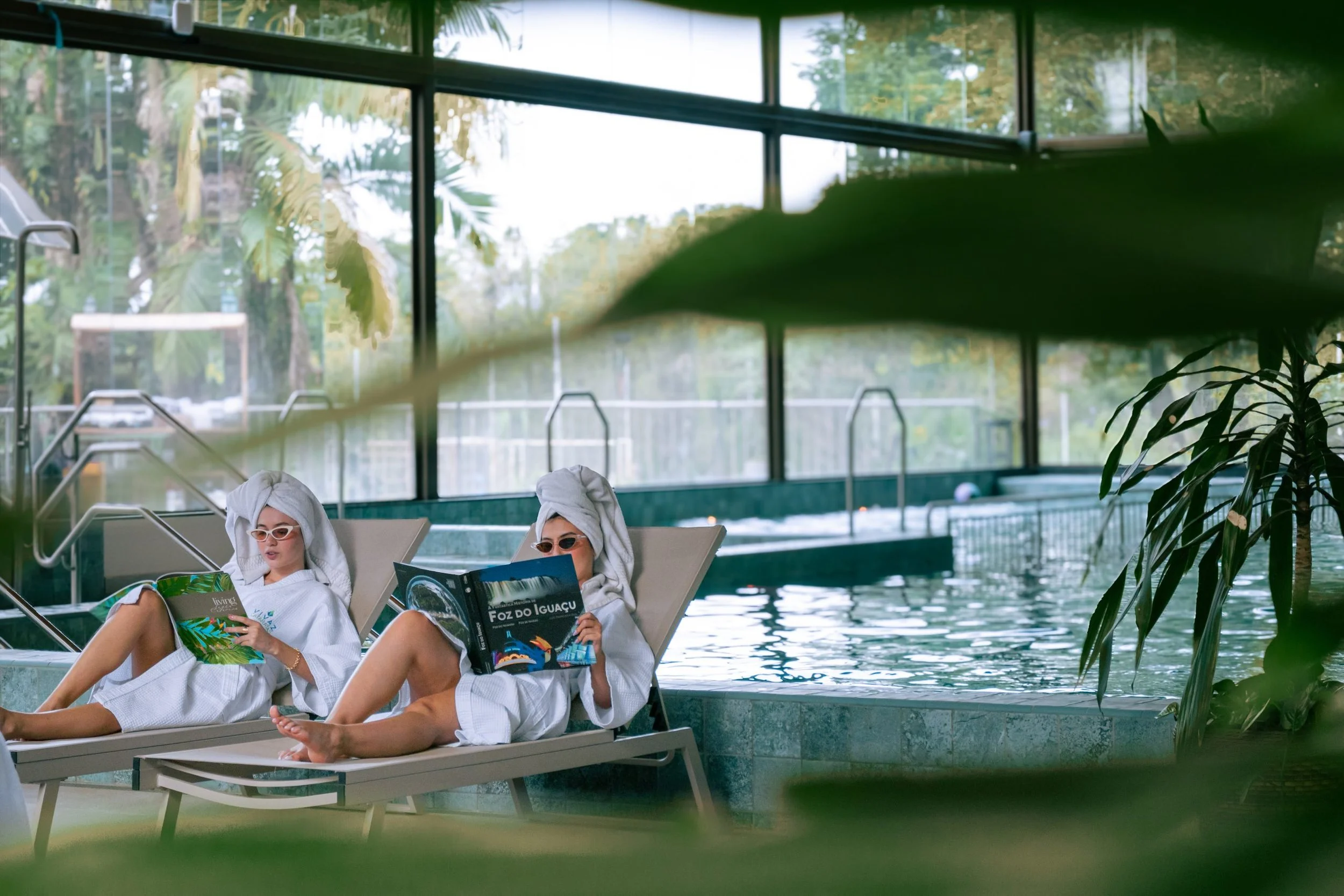 Two women in white robes and towels on their heads are lounging by an indoor swimming pool, reading magazines, with large windows showing outdoor trees and sky.