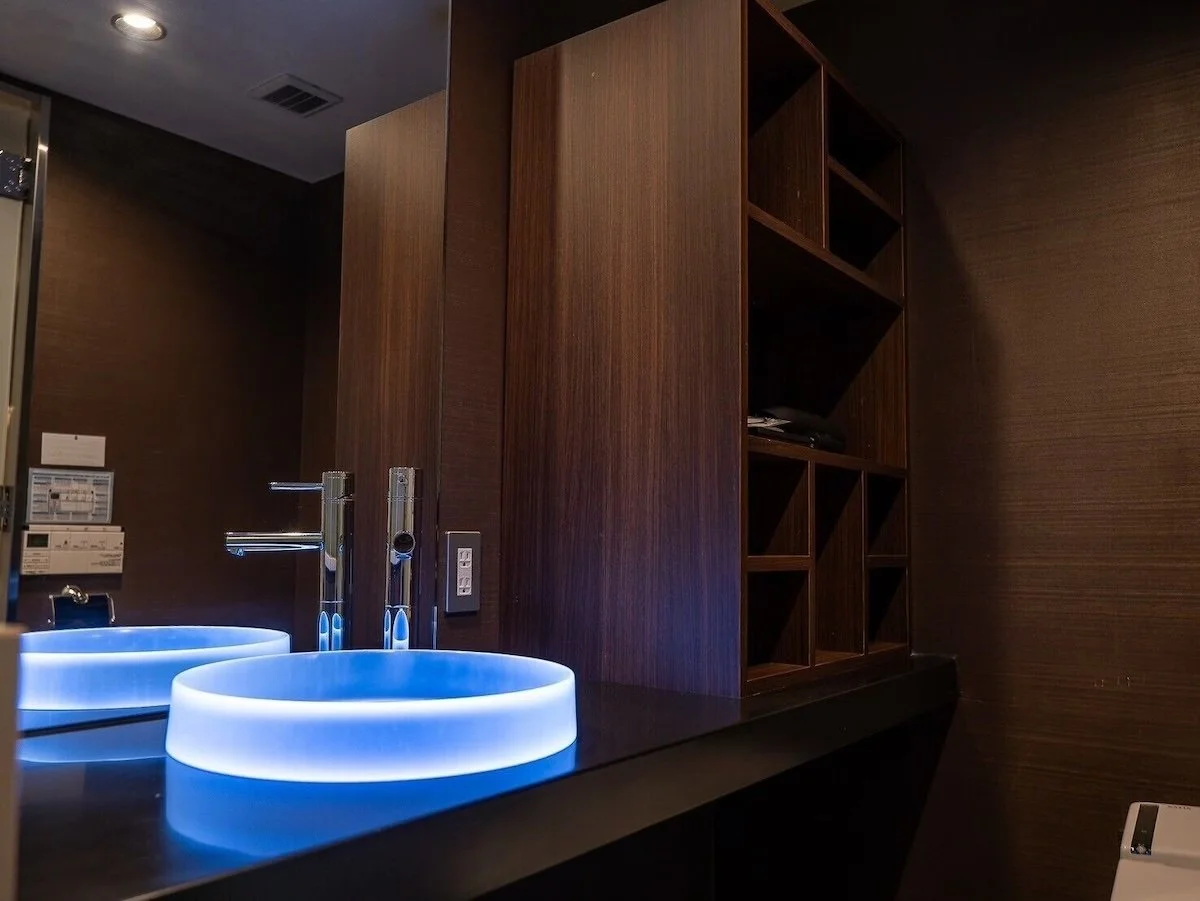 Modern bathroom with illuminated oval sink, wooden shelving unit, and dark countertop.