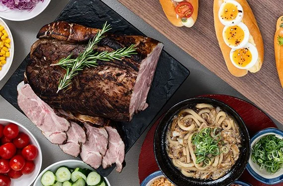 A roasted beef tenderloin with rosemary on a black cutting board, a bowl of cherry tomatoes, a bowl of cucumber slices, a bowl of ramen with green onions, and slices of toasted bread topped with soft-boiled eggs and herbs.