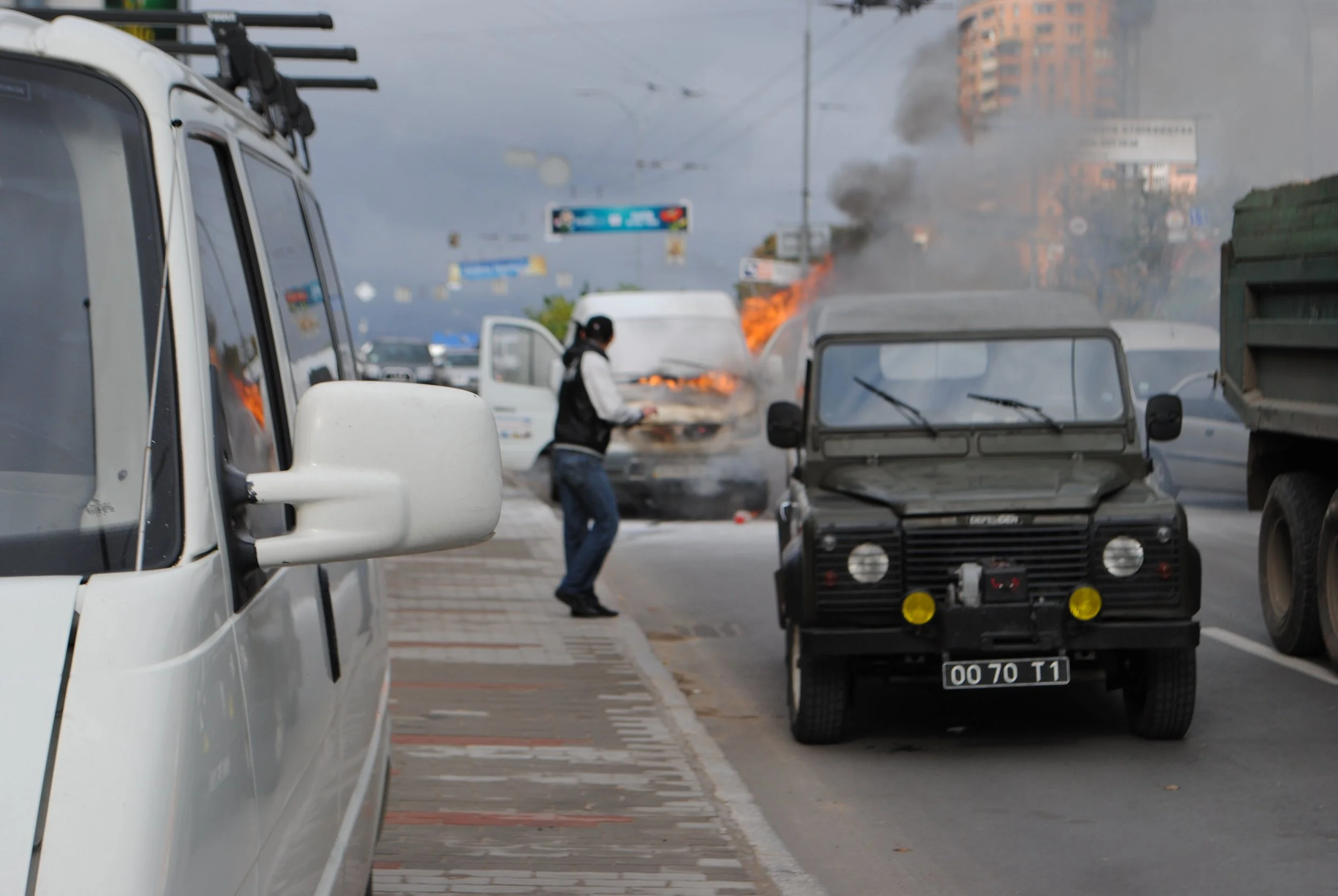 A car at the side of a busy street with a fire and black smoke coming from the back of another vehicle. A person is walking nearby, and other cars are parked along the road. Kiev November 2011, Marina Parrish ©