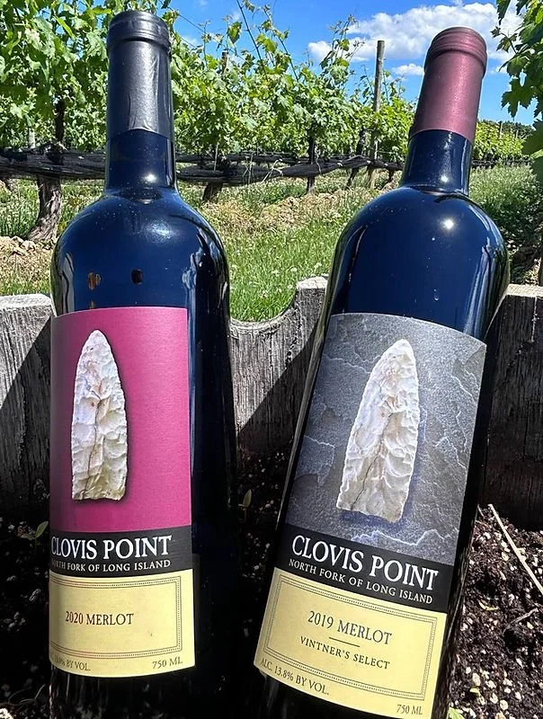 Two bottles of Clovis Point Merlot wine, one from 2020 and the other from 2019, outdoors against a vineyard background.