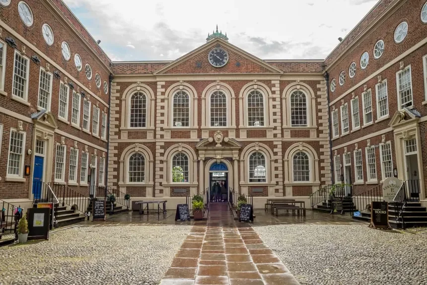 Historic brick building with large arched windows and a central clock, surrounded by a courtyard with cobblestone and brick pathway, outdoor benches, and chalkboard signs.
