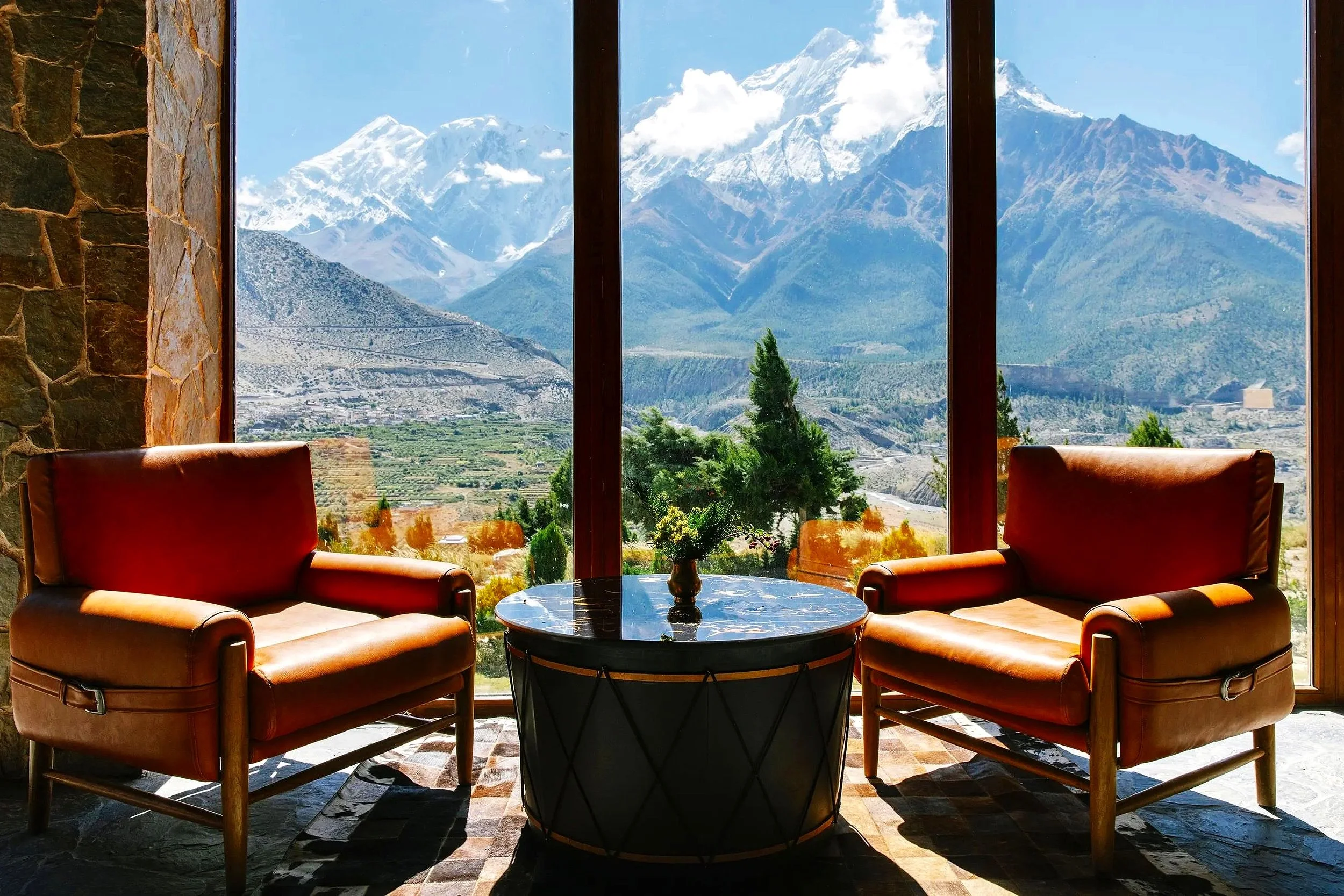 Living room with two leather armchairs facing a large window overlooking snow-capped mountains, green trees, and a valley.