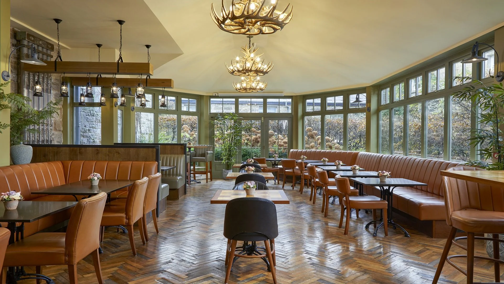 A cozy, well-lit restaurant with large windows showing a garden view, featuring wooden floors, brown leather chairs, and small flower arrangements on tables, with antler chandeliers hanging from the ceiling.