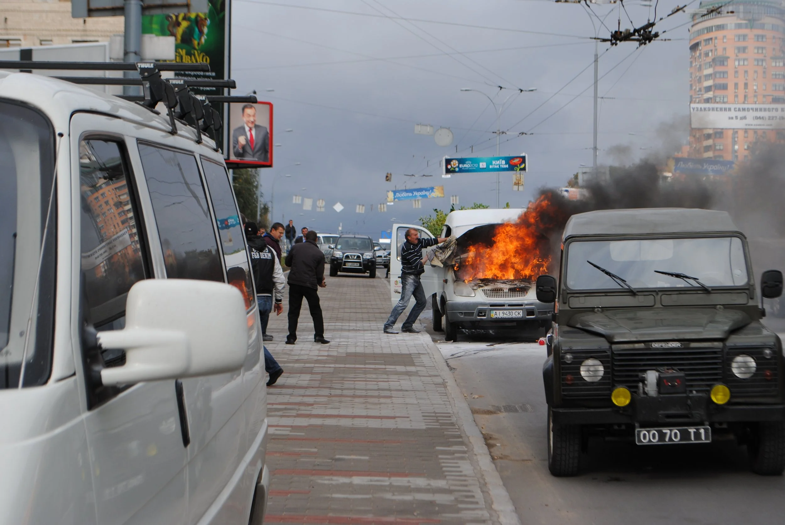 A crowd of people standing and walking near cars on a city street; there is a police or emergency vehicle with flashing lights and some dark smoke and flames coming from a vehicle. In the background, there are tall buildings, billboards, and electric