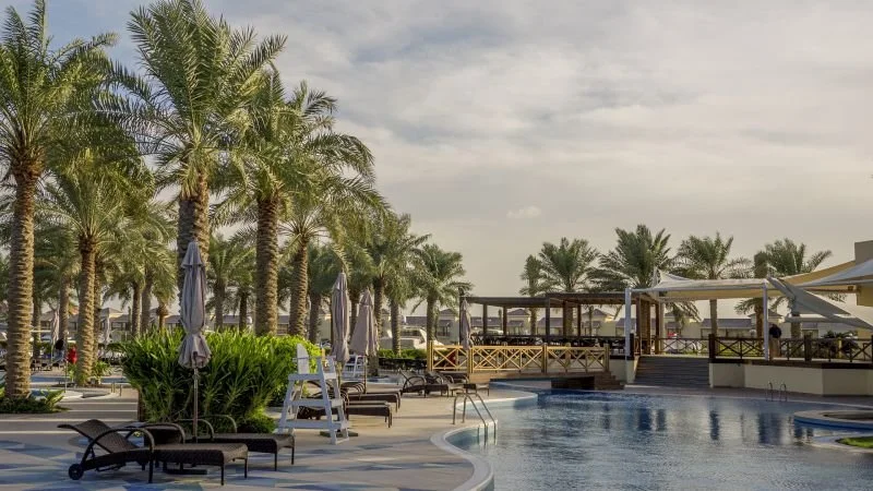 Poolside area at a tropical resort with palm trees, lounge chairs, shaded cabanas, and an outdoor bar or restaurant