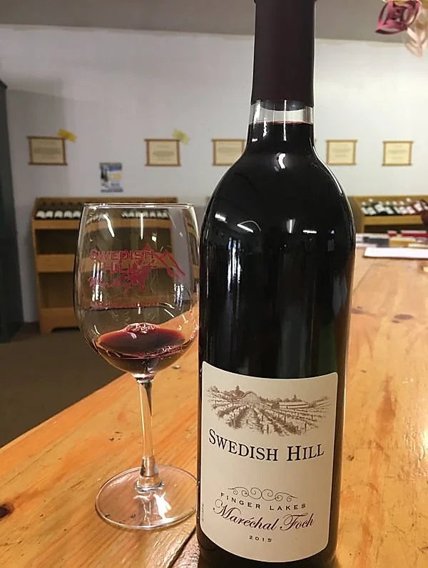 A bottle of Swedish Hill Marechal Foch red wine from 2015 and a glass of red wine on a wooden table in a wine shop.