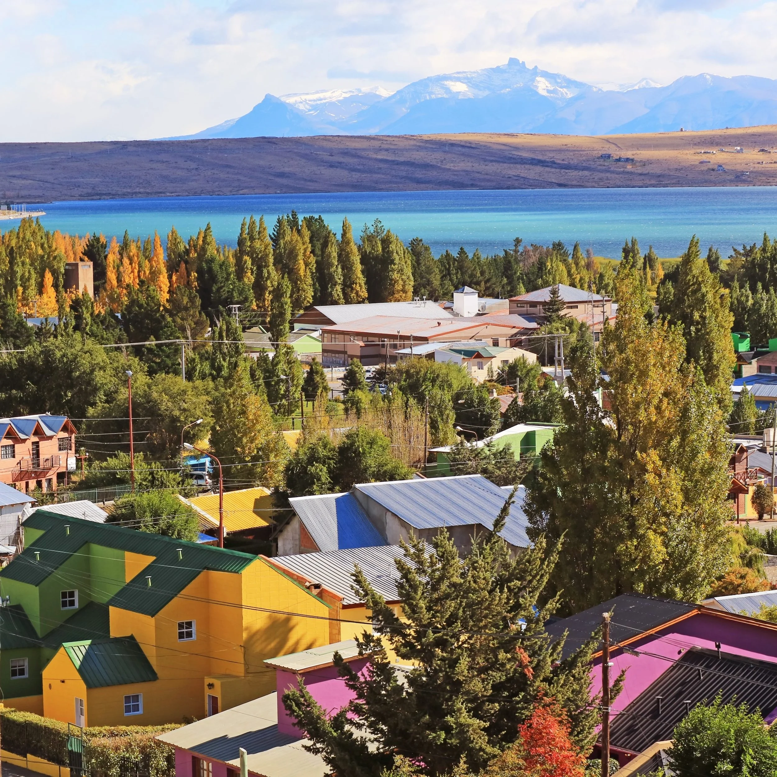 Colorful houses with brightly painted walls and metal roofs in a neighborhood with trees and power lines, overlooking a lake with a mountain range in the background.