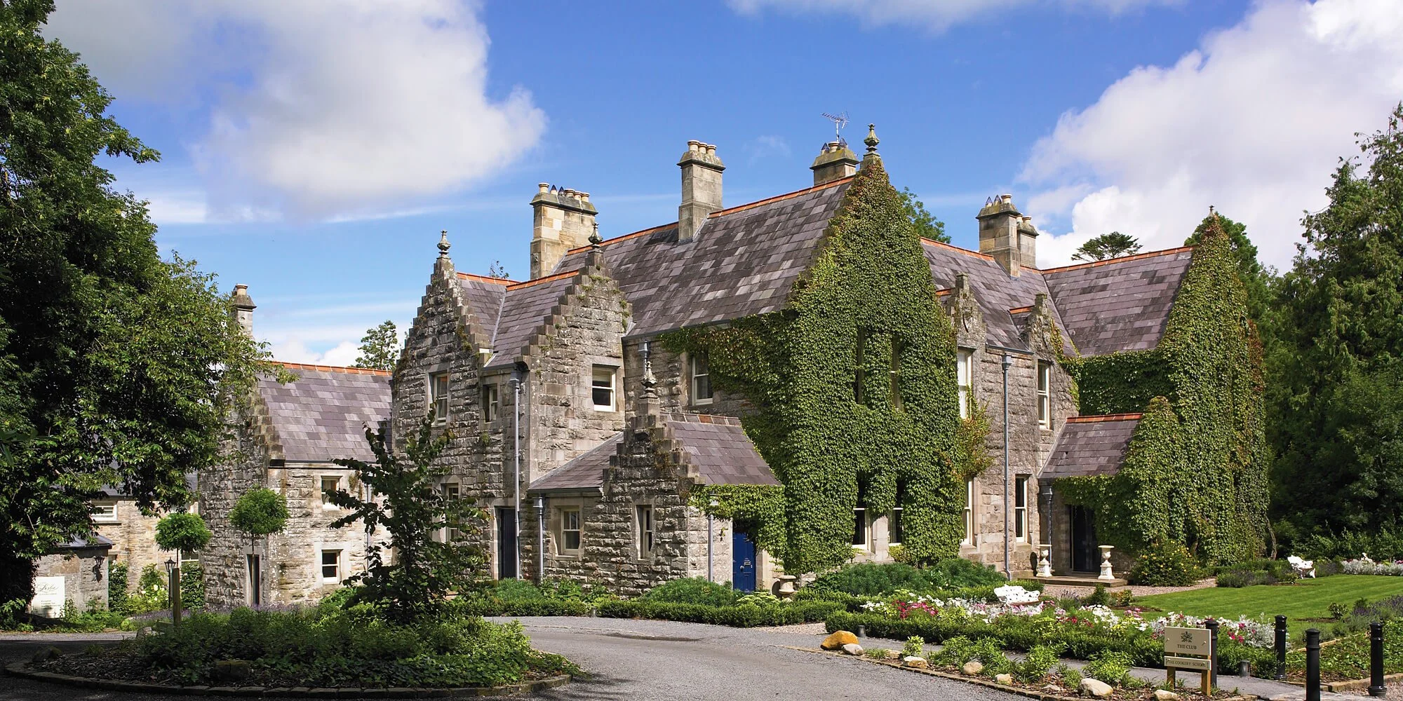 Stone building with ivy-covered walls, multiple chimneys, surrounded by greenery under a blue sky with clouds.