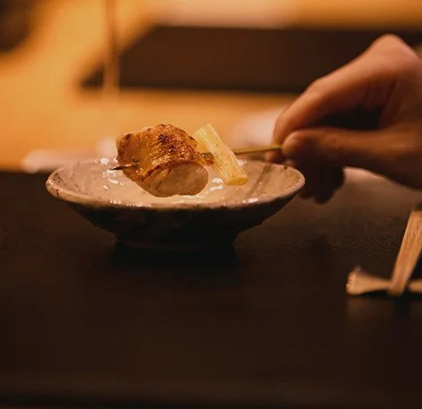 A hand holds a skewer with grilled chicken and pineapple over a small ceramic dish.