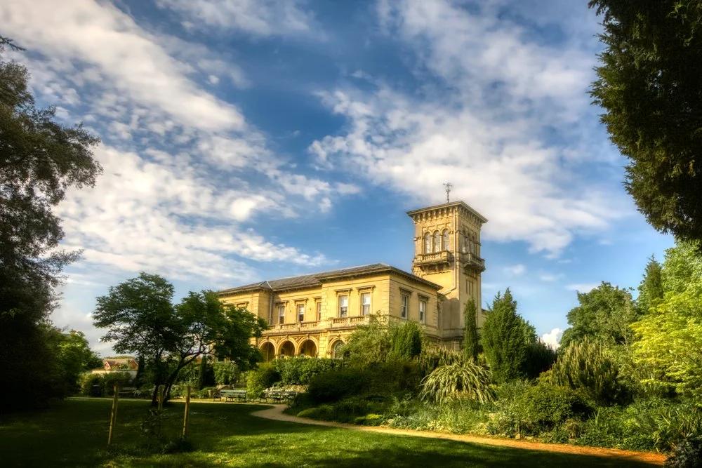 A historic building with ornate architecture, surrounded by greenery and trees, under a partly cloudy sky.