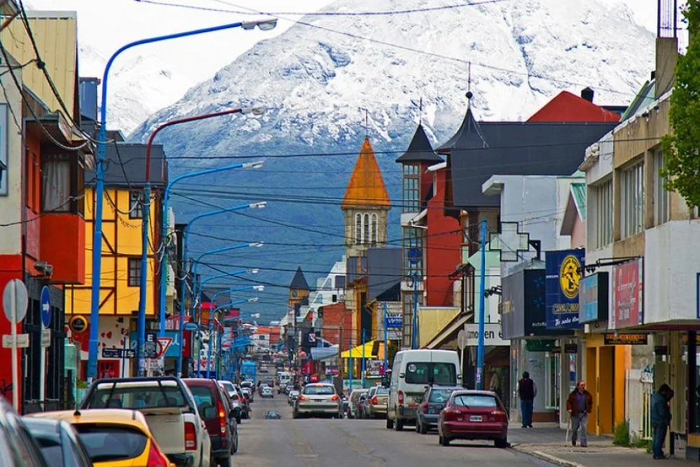 Street view in a town with colorful buildings, parked cars, and pedestrians. Mountains are visible in the background.