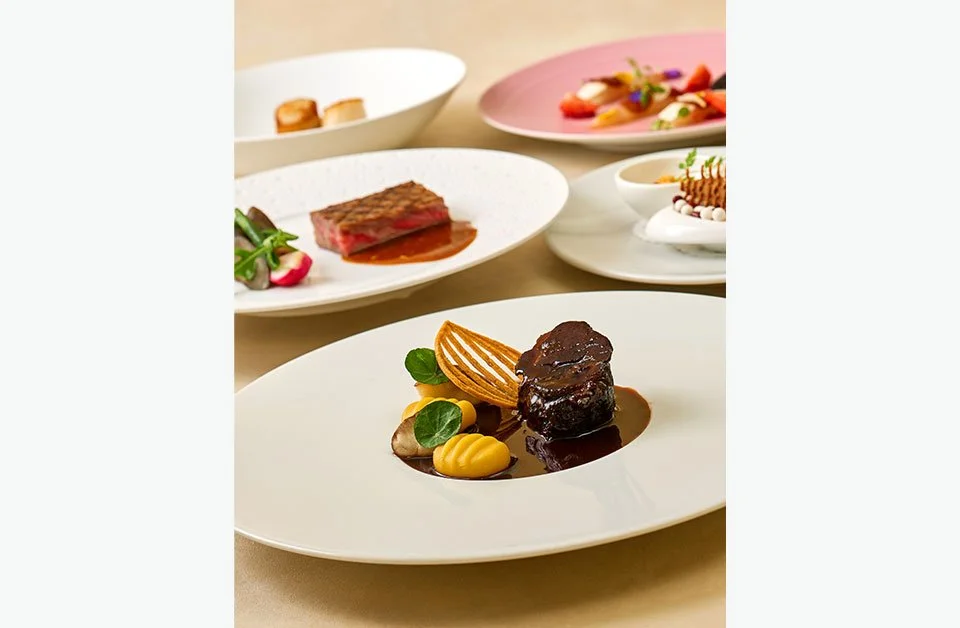 Elegant display of multiple gourmet dishes on white plates, including a main course with a meat chop, vegetables, and decorative elements, set on a beige tablecloth.