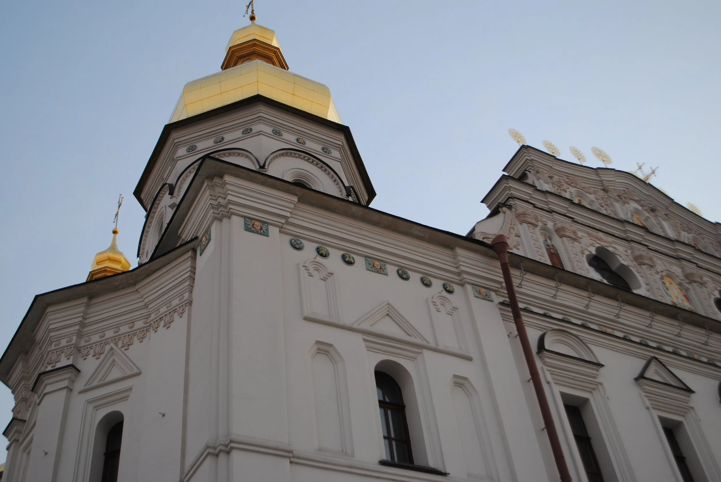 A church with a tall, gold-domed tower and intricate white facade, featuring decorative details and small paintings along the trim, seen against a light blue sky.
