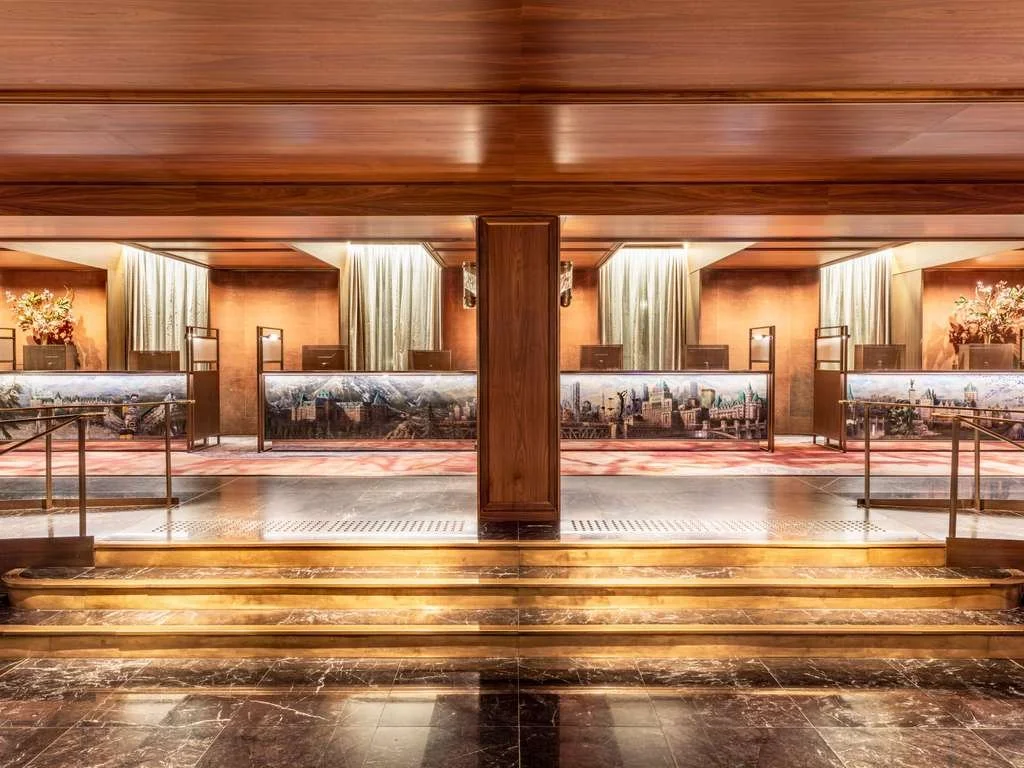 Hotel reception desk with several counters, gold accents, marble floors, and view of city skyline murals behind the counters.