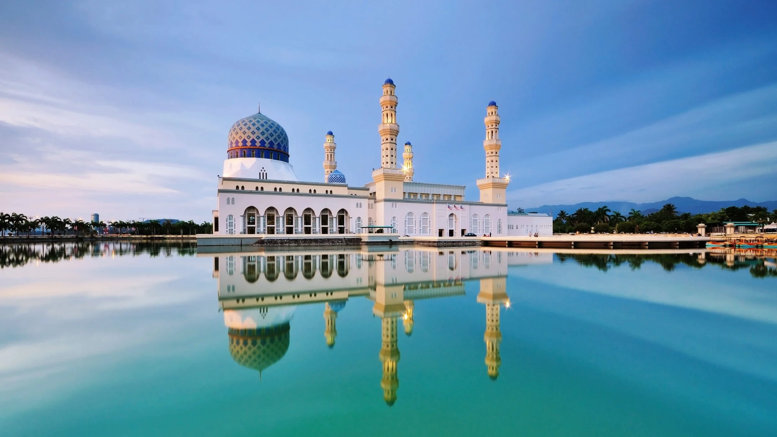 A large mosque with a blue dome and four tall minarets, reflected in a calm body of water with a cityscape and mountains in the background at dusk.