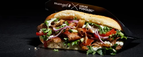 Close-up of a doner kebab sandwich with sliced meat, fresh vegetables, and sauce in a bread roll, wrapped in black paper.