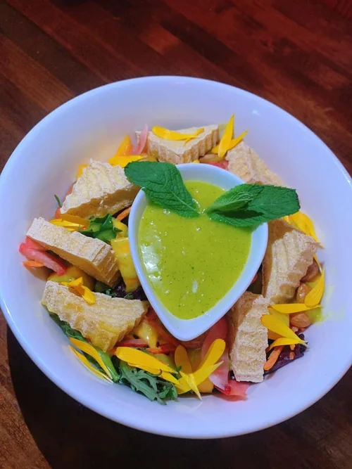 A bowl of salad with pieces of fried bread, colorful vegetables, and edible flower petals, topped with a small cup of green dressing or sauce and garnished with two large green mint leaves.