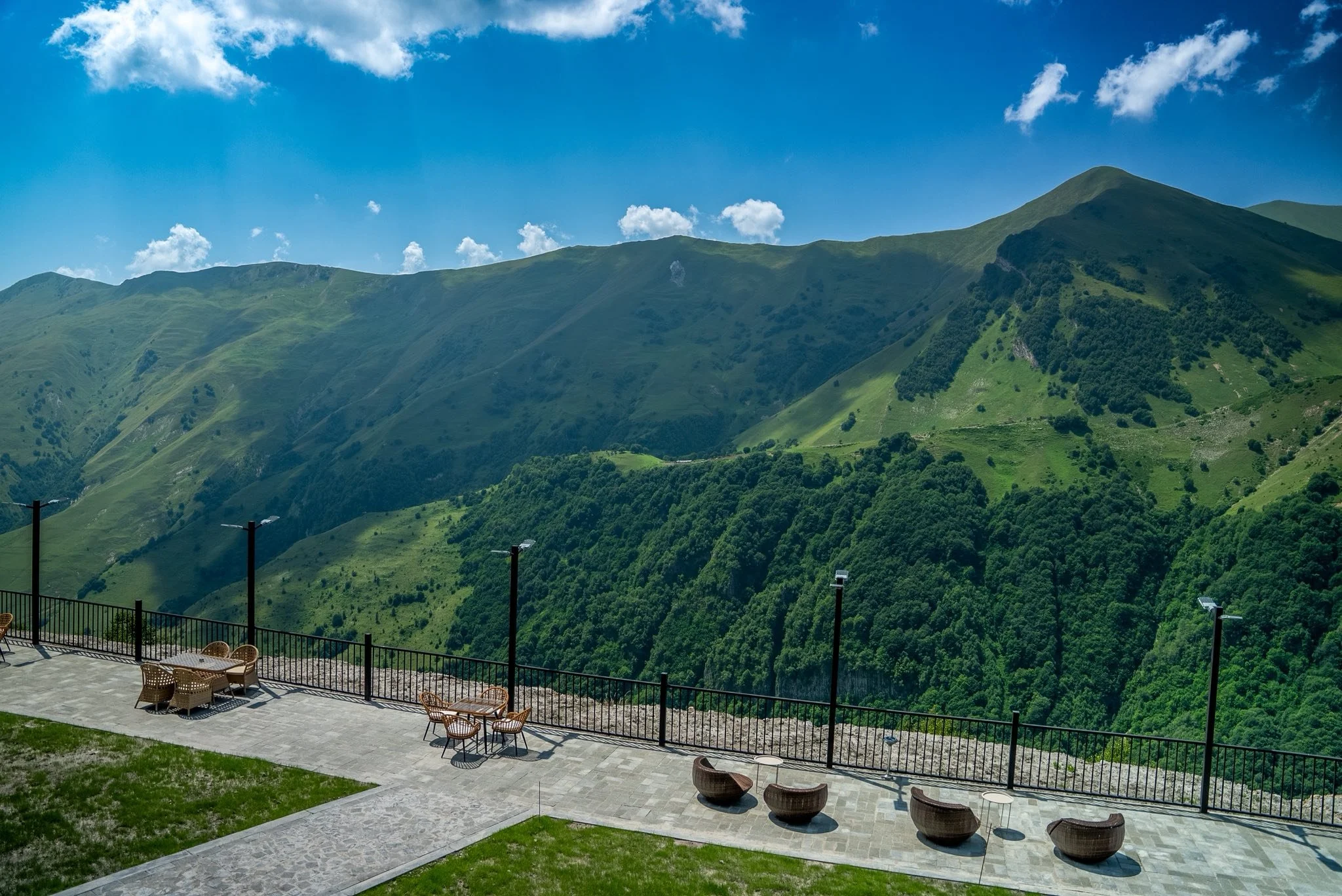 An outdoor terrace with chairs and tables overlooks lush green mountains under a blue sky with clouds.