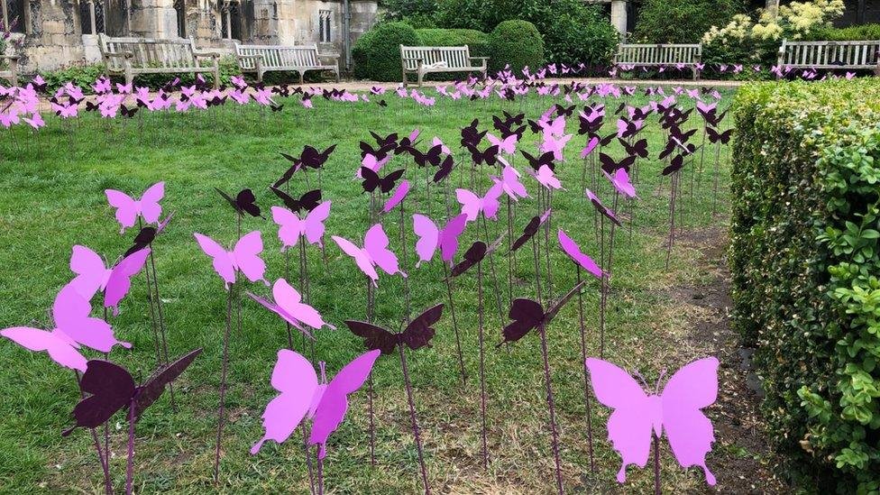 An outdoor art installation featuring numerous purple and black butterfly-shaped cutouts on sticks arranged in rows on a grassy area in a park, with benches and trimmed bushes in the background.