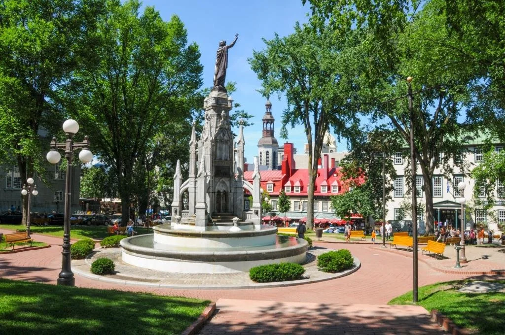 A park with a fountain featuring a statue of a man with one arm raised, surrounded by trees, benches, and lamp posts, with buildings in the background.