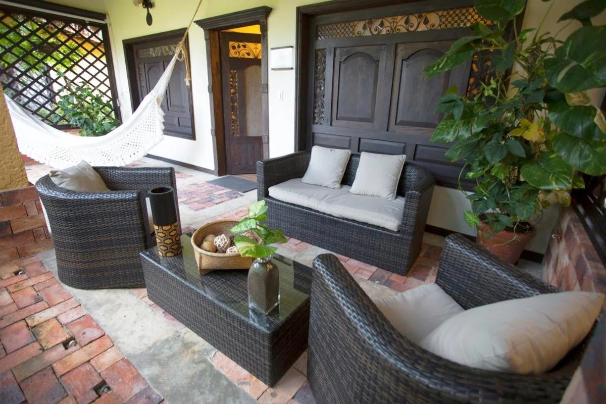 Outdoor patio with wicker furniture including a loveseat with beige cushions, two armchairs with beige cushions, a glass-top coffee table with a vase containing green leaves, a basket with decorative balls, a large potted plant, a hammock, brick and 
