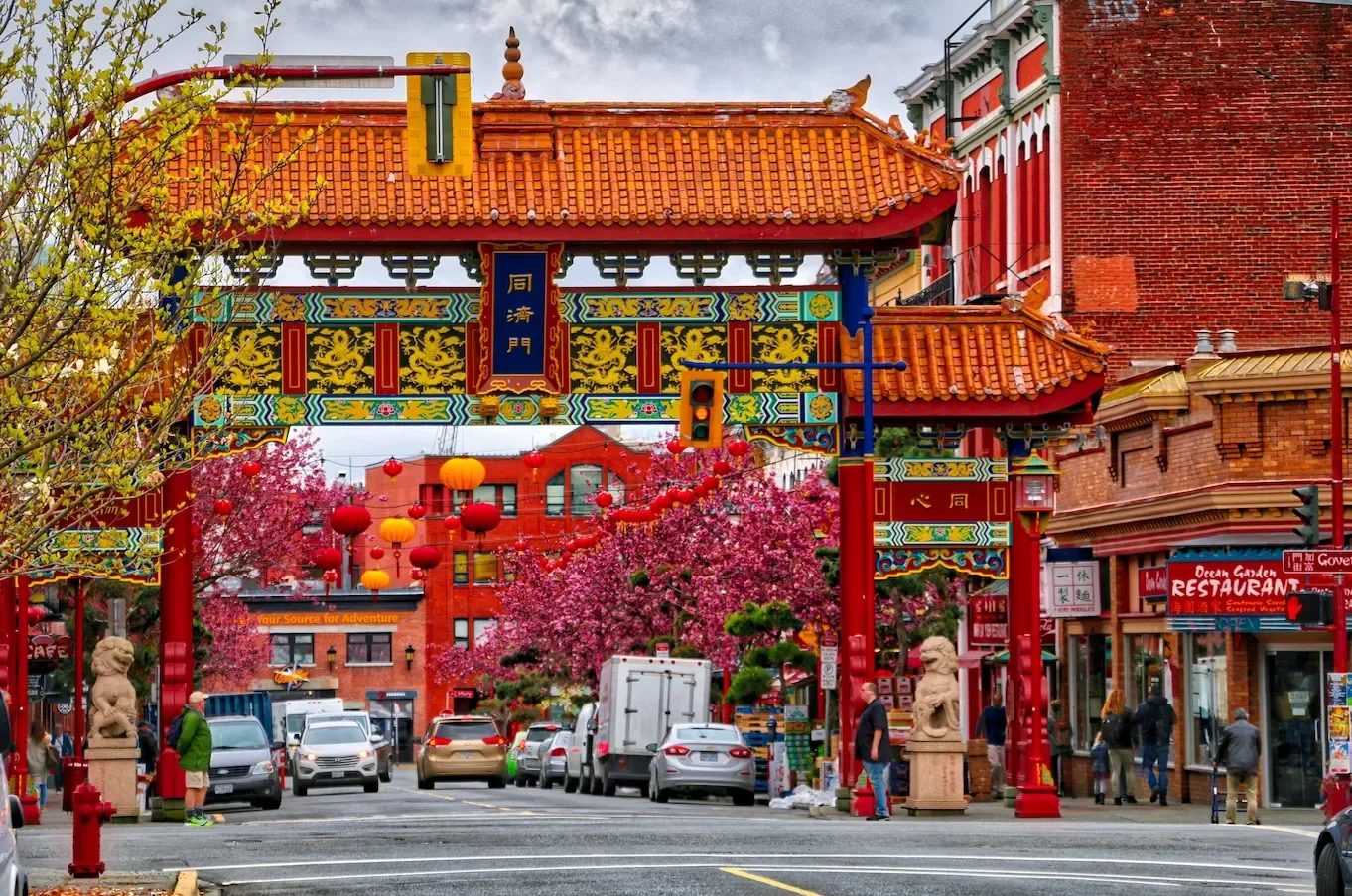 A busy street scene in Chinatown with an ornate Chinese arch gate, red lanterns, pink blooming trees, cars, and pedestrians.
