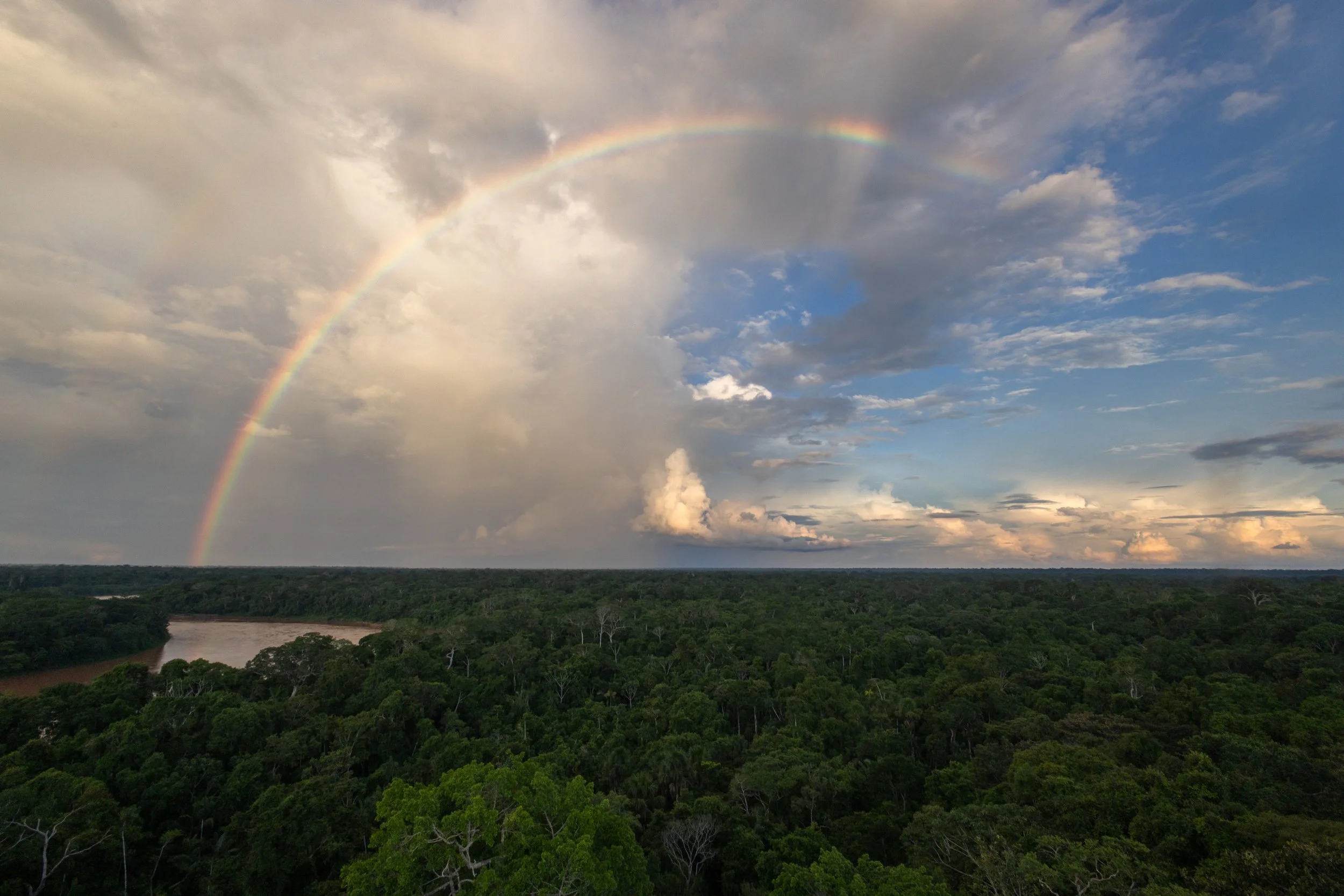 A rainbow arches across a partly cloudy sky over a landscape of dense green forest and a river in the distance.