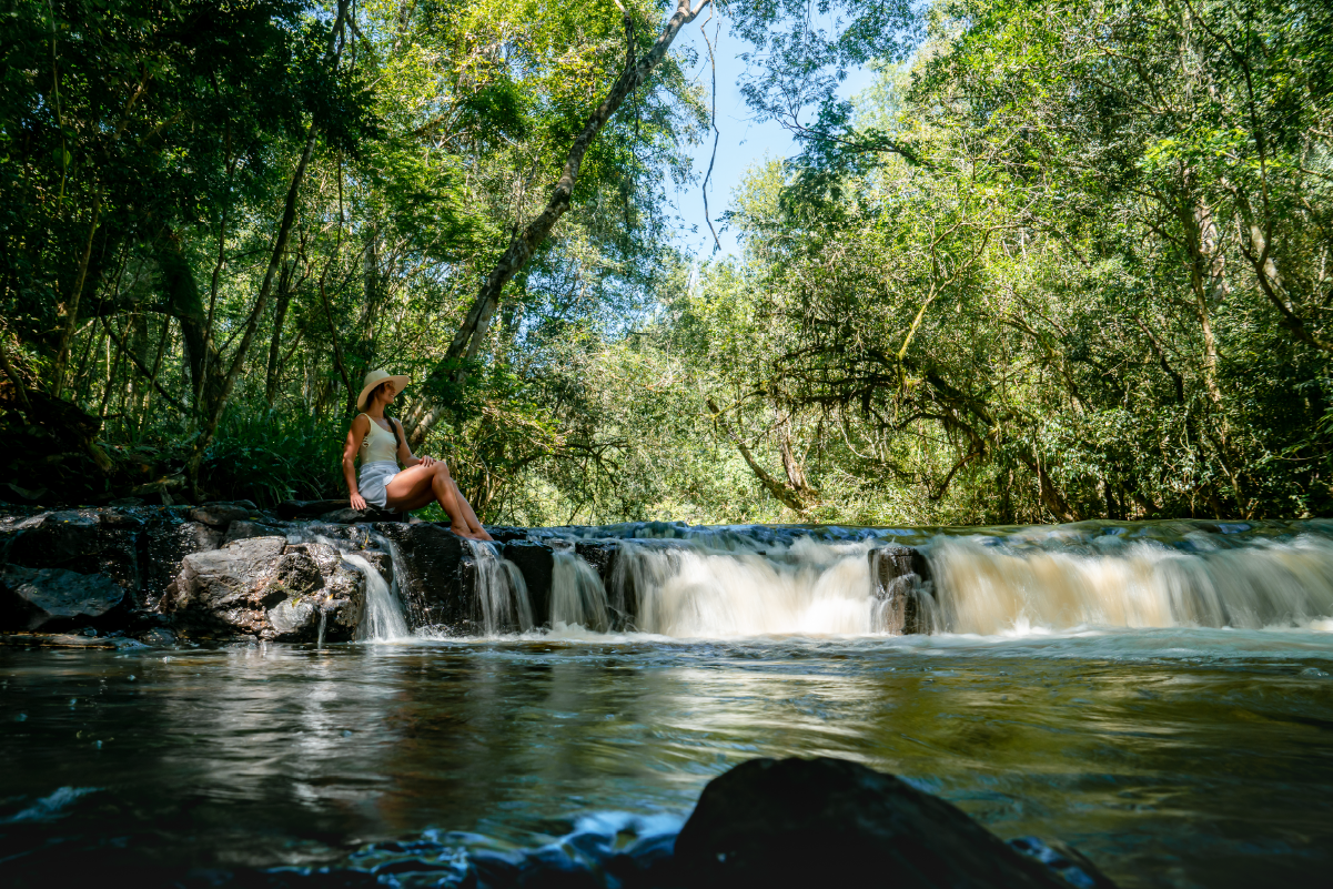 A woman in a light-colored hat and dress is sitting on rocks next to a flowing river in a lush, green forest with a blue sky visible through the canopy.