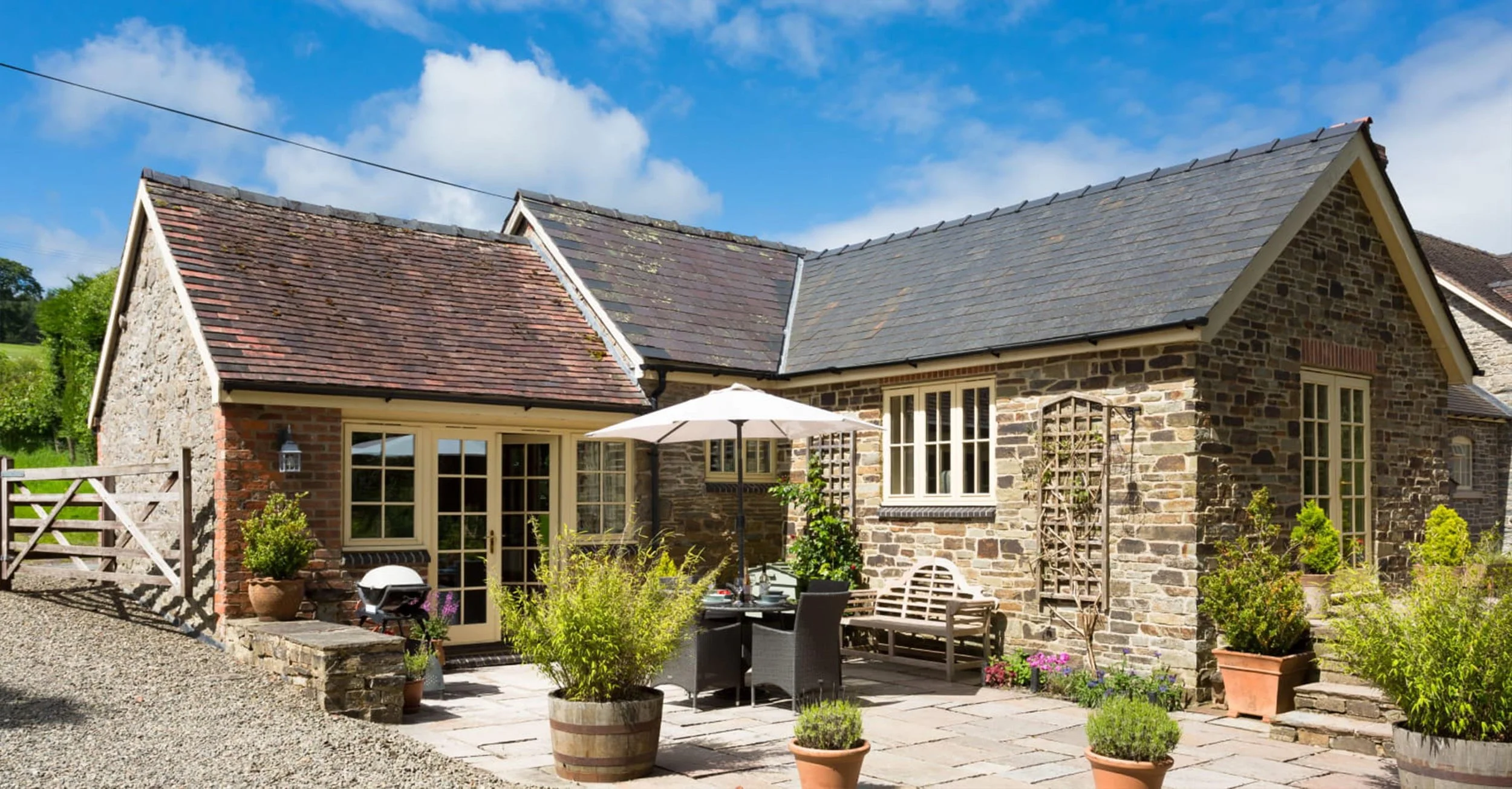 A stone house with a patio, outdoor table with an umbrella, potted plants, and a gravel pathway under a blue sky with clouds.