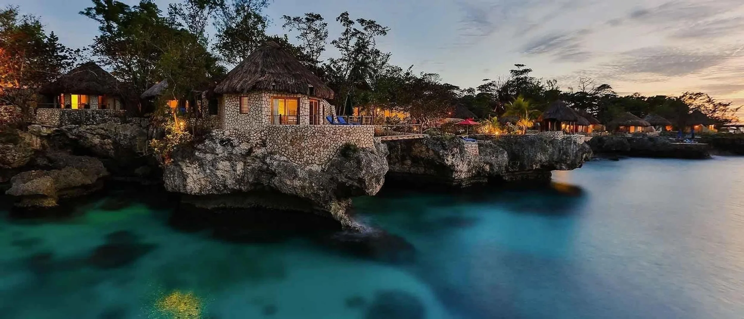 Beachfront cottages built on rocky cliffs overlooking turquoise water at sunset with trees and a cloudy sky in the background.